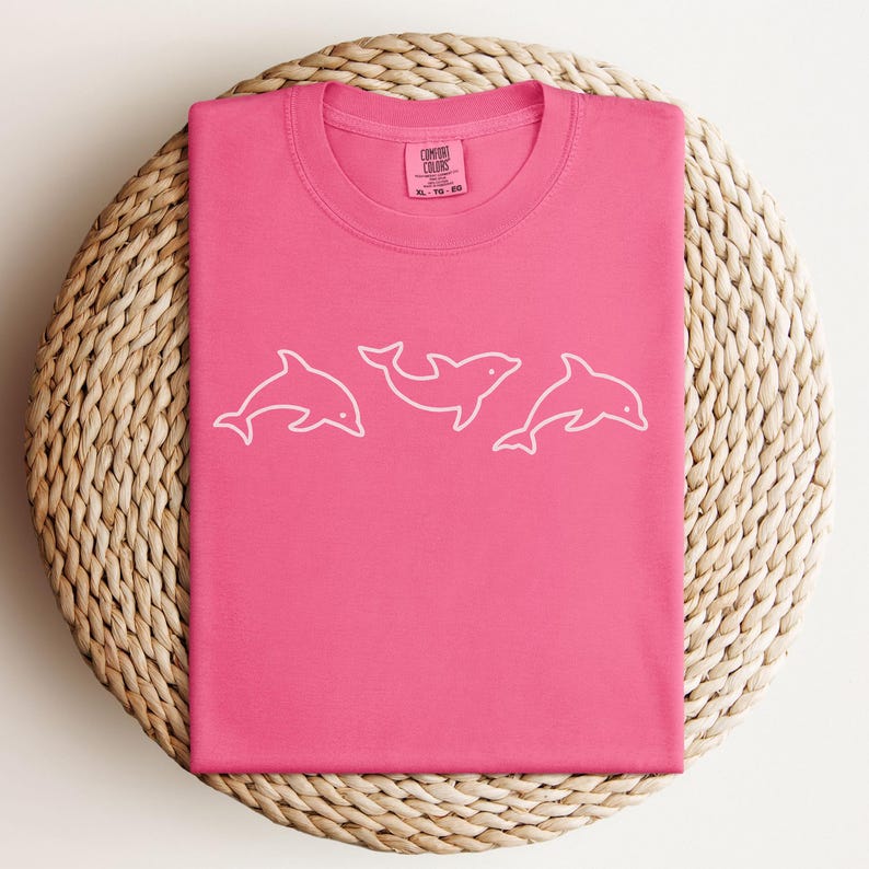May include: A hot pink Comfort Colors t-shirt featuring a white outline of three dolphins. The shirt is folded neatly on a woven, circular placemat. The Comfort Colors label is visible at the neckline.