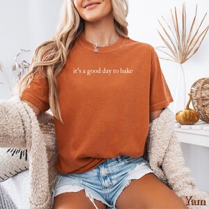 Puede incluir: Camiseta naranja quemado con la frase "it's a good day to bake" en letras blancas. La camiseta se combina con pantalones cortos de mezclilla y una rebeca color crema. El conjunto sugiere un estilo casual y c&oacute;modo.