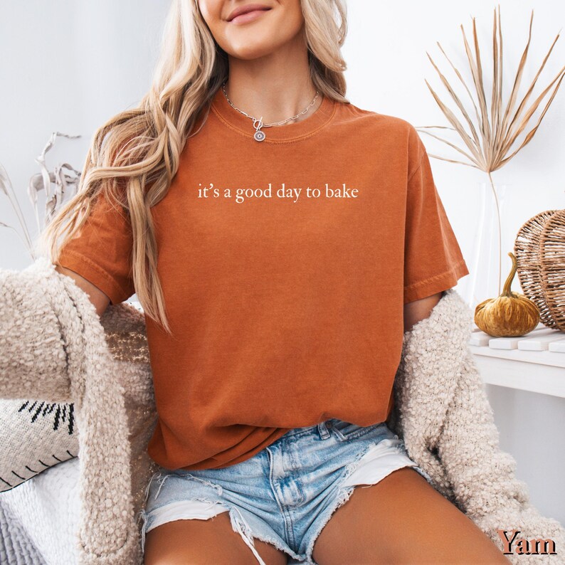 Puede incluir: Camiseta naranja quemado con la frase "it's a good day to bake" en letras blancas. La camiseta se combina con pantalones cortos de mezclilla y una rebeca color crema. El conjunto sugiere un estilo casual y c&oacute;modo.