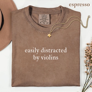 May include: An espresso-colored t-shirt with a crew neckline and the white text "easily distracted by violins" printed on the front. The shirt features a Comfort Colors brand label at the inner neck, size XL, and is made of 100% cotton fabric.