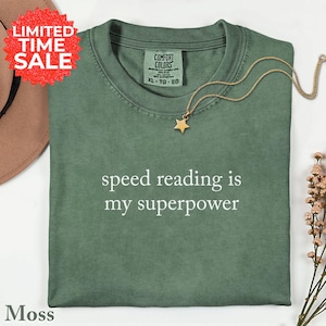 May include: A moss green t-shirt with the text "speed reading is my superpower" in white. A gold star pendant necklace rests on the shirt. The shirt is folded, and a "Limited Time Sale" sticker is visible.