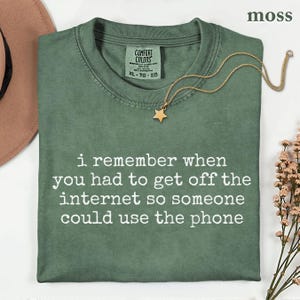 May include: A moss green short-sleeve t-shirt with a crew neckline and the white printed text: i remember when you had to get off the internet so someone could use the phone. The shirt features a Comfort Colors brand label and a gold star necklace.