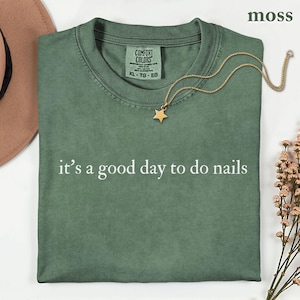 May include: A moss green crew neck t-shirt with the white text "it's a good day to do nails" printed on the front. The shirt features a Comfort Colors brand label at the inner neckline and is displayed with a gold star pendant necklace.