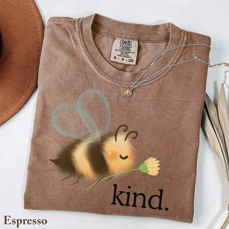 May include: Espresso-colored t-shirt with a graphic of a cartoon bee holding a flower, with the word "kind" printed below. The shirt has a crew neck and short sleeves. A silver necklace with a small pendant is draped across the shirt.