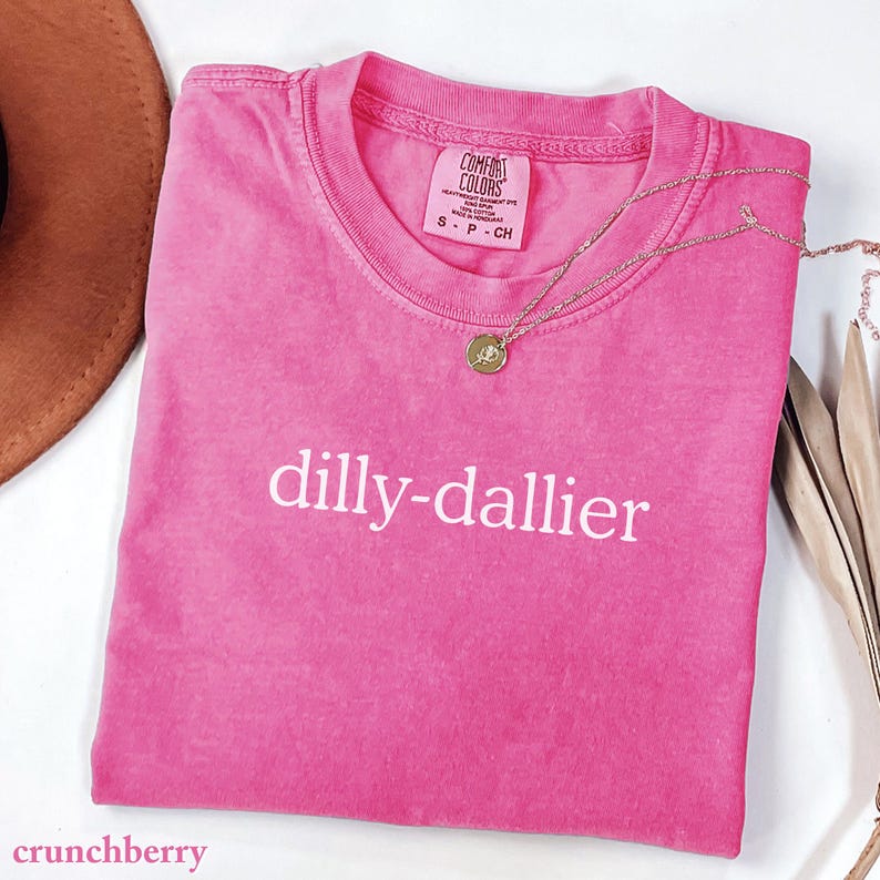 May include: A pink t-shirt with the word "dilly-dallier" printed in white. The shirt has a crew neck and short sleeves. A gold necklace with a pendant is draped across the shirt. The shirt is on a white surface.