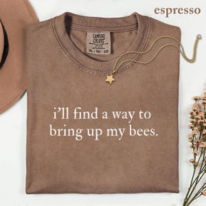 May include: An espresso-colored crew neck t-shirt with the white text "i'll find a way to bring up my bees." printed on the front. The shirt features a Comfort Colors brand label at the inner neckline and is size XL.