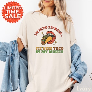 May include: Ivory-colored t-shirt with the text "I'M INTO FITNESS... FIT'NESS TACO IN MY MOUTH" and a graphic of a taco in a mouth. The shirt has rolled-up sleeves and is paired with a denim jacket. A red and orange "LIMITED TIME SALE" sticker is in the upper left corner.