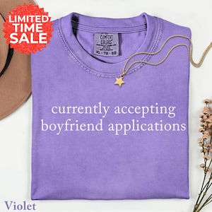 May include: A violet-colored t-shirt with the text "currently accepting boyfriend applications" in white. A gold star pendant necklace rests on the shirt. The shirt has a Comfort Colors tag and a "Limited Time Sale" sticker.