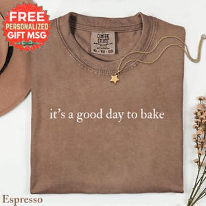 May include: Espresso-colored t-shirt with the phrase "it's a good day to bake" in white. A gold star pendant necklace rests on the shirt. The shirt has a Comfort Colors tag, size XL-TG-EG. A "FREE PERSONALIZED GIFT MSG" sticker is in the upper left.