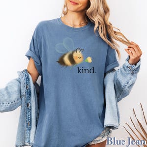 May include: Blue jean colored t-shirt featuring a cartoon bee holding a yellow flower. The bee has black and yellow stripes and transparent wings. The word "kind" is printed in black below the bee. The shirt has rolled-up sleeves.