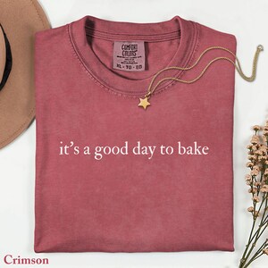 Puede incluir: Camiseta carmes&iacute; doblada con la frase blanca &laquo;it's a good day to bake&raquo;. Un collar con colgante de estrella dorada descansa sobre la camiseta. La camiseta tiene una etiqueta Comfort Colors.