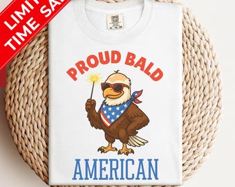 May include: A white t-shirt featuring a graphic of a bald eagle wearing sunglasses and a star-spangled bandana, holding a sparkler. The text PROUD BALD is arched above in red, and the word AMERICAN is printed below in blue. The shirt has a Comfort Colors label.