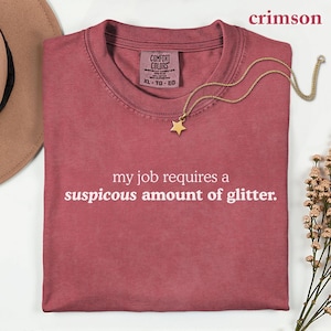 May include: A crimson short-sleeved t-shirt featuring the white printed text 'my job requires a suspicous amount of glitter.' The shirt has a Comfort Colors brand label at the inner neckline and is paired with a gold-toned star pendant necklace.
