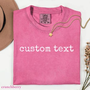 May include: A folded, pink t-shirt with the words "custom text" printed in white. A gold-colored star pendant necklace rests on the shirt. The shirt has a Comfort Colors tag. A brown hat and sprig of flowers are in the background.