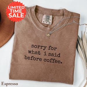 May include: Espresso-colored Comfort Colors t-shirt with the text "sorry for what I said before coffee." A silver necklace with a small pendant is draped on the shirt. A red "LIMITED TIME SALE" sticker is in the upper left corner.