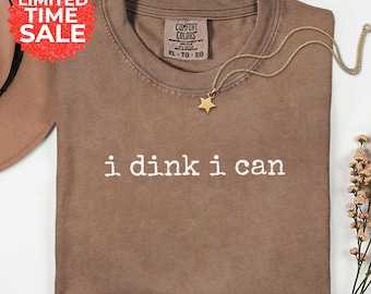 May include: A brown short-sleeved t-shirt with the white text i dink i can printed on the front. The shirt features a crew neckline and a Comfort Colors brand label inside the collar. A gold-toned necklace with a small star charm rests on the shirt.