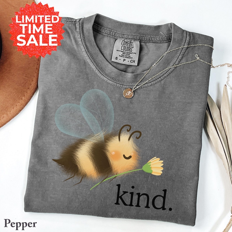 May include: A gray Comfort Colors t-shirt featuring a cartoon bee holding a flower, with the word "kind" printed below. The shirt has a crew neck and short sleeves. A silver necklace with a small pendant is draped across the shirt.