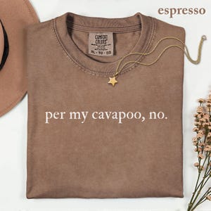 May include: An espresso-colored t-shirt with a crew neckline and the text 'per my cavapoo, no.' printed in white. The shirt features a Comfort Colors label at the inner neck and is displayed with a gold-toned star pendant necklace.