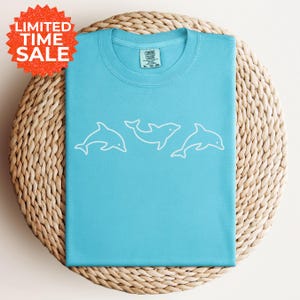 May include: A light blue t-shirt featuring three white outline drawings of dolphins. The shirt has a crew neck and short sleeves. A red and white "LIMITED TIME SALE" sticker is in the upper left corner.