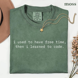 May include: A moss green crew neck t-shirt with the white text "i used to have free time, then i learned to code." The shirt features a Comfort Colors label at the inner neckline and is displayed with a gold star pendant necklace.