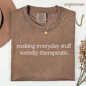 May include: An espresso-colored crew neck t-shirt with the white text "making everyday stuff weirdly therapeutic." printed on the front. The shirt features a Comfort Colors label at the inner neckline and is paired with a gold star pendant necklace.