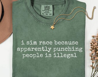 May include: A moss green short-sleeve t-shirt with a crew neckline and the white printed text: i sim race because apparently punching people is illegal. The shirt features a Comfort Colors brand label at the inner neck and a gold star necklace.