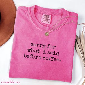 May include: A bright pink short-sleeved t-shirt with a crew neckline and a Comfort Colors brand label. The shirt features the black printed text, sorry for what i said before coffee. A delicate gold-toned necklace with a small circular pendant rests on the shirt.