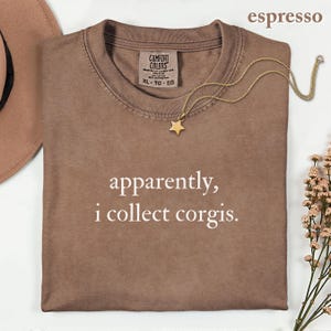 May include: An espresso-colored t-shirt with a crew neckline and the white text "apparently, i collect corgis." printed on the front. The shirt features a Comfort Colors brand label at the inner neck and is displayed with a gold star pendant necklace.