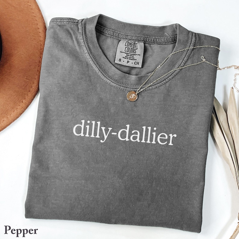 May include: A gray Comfort Colors t-shirt with the word "dilly-dallier" printed in white. A silver necklace with a small pendant rests on the shirt. The shirt is folded and displayed with a brown hat and decorative leaves.