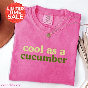 May include: A pink t-shirt with the phrase "cool as a cucumber" in green lettering. The shirt has a crew neck and short sleeves. A gold necklace with a pendant rests on the shirt. A red "Limited Time Sale" sticker is in the upper left corner.