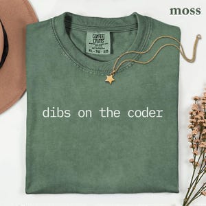 May include: A moss green short-sleeve t-shirt with the text "dibs on the coder" printed in white across the chest. The shirt features a Comfort Colors brand label at the inner neckline, a gold-toned chain necklace with a star pendant, and a hat.