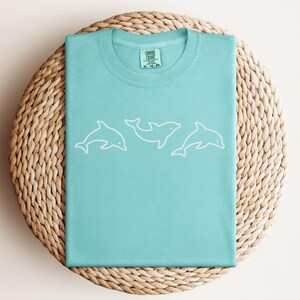 May include: A light blue t-shirt with a white outline of three dolphins. The shirt has a crew neck and short sleeves. The tag on the shirt reads "Comfort Colors" and includes size information.