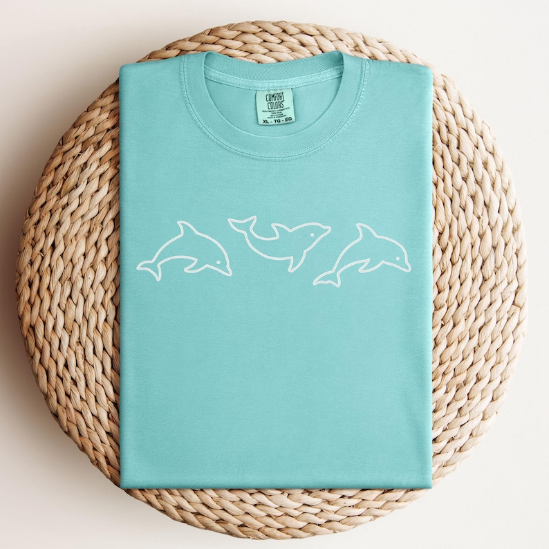 May include: A light blue t-shirt with a white outline of three dolphins. The shirt has a crew neck and short sleeves. The tag on the shirt reads "Comfort Colors" and includes size information.