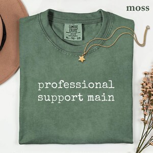 May include: A moss green crew neck t-shirt with the text professional support main printed in white on the chest. The shirt features a Comfort Colors label at the inner neckline with size XL-TG-EG. A gold star pendant necklace rests on the fabric.