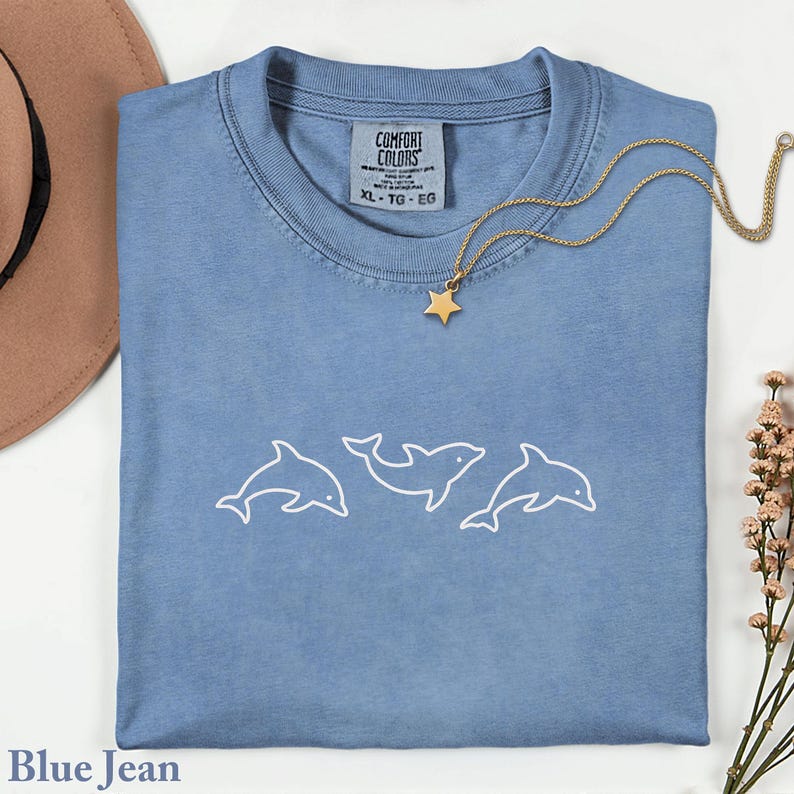 May include: A blue jean colored t-shirt with a white outline of three dolphins. The shirt has a Comfort Colors tag and a gold necklace with a star pendant. A brown hat and flowers are in the background.