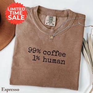 May include: Espresso-colored t-shirt with the text "99% coffee 1% human" in black. The shirt has a crew neck and short sleeves. A silver necklace with a small pendant is draped across the shirt. A "Limited Time Sale" sticker is in the upper left corner.