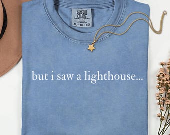 May include: A blue jean colored t-shirt with the text but i saw a lighthouse printed in white across the chest. The shirt features a Comfort Colors label at the inner neckline and is displayed with a gold star pendant necklace.