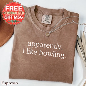 May include: Espresso-colored t-shirt with the text "apparently, i like bowling." A silver necklace with a pendant rests on the shirt. The shirt has a Comfort Colors tag. A red and green gift message is in the upper left.
