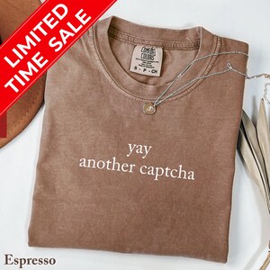 May include: Espresso-colored t-shirt with the text "yay another captcha" in white. A silver necklace with a small pendant rests on the shirt. The shirt has a crew neck and a tag that reads "Comfort Colors". A red banner reads "LIMITED TIME SALE".