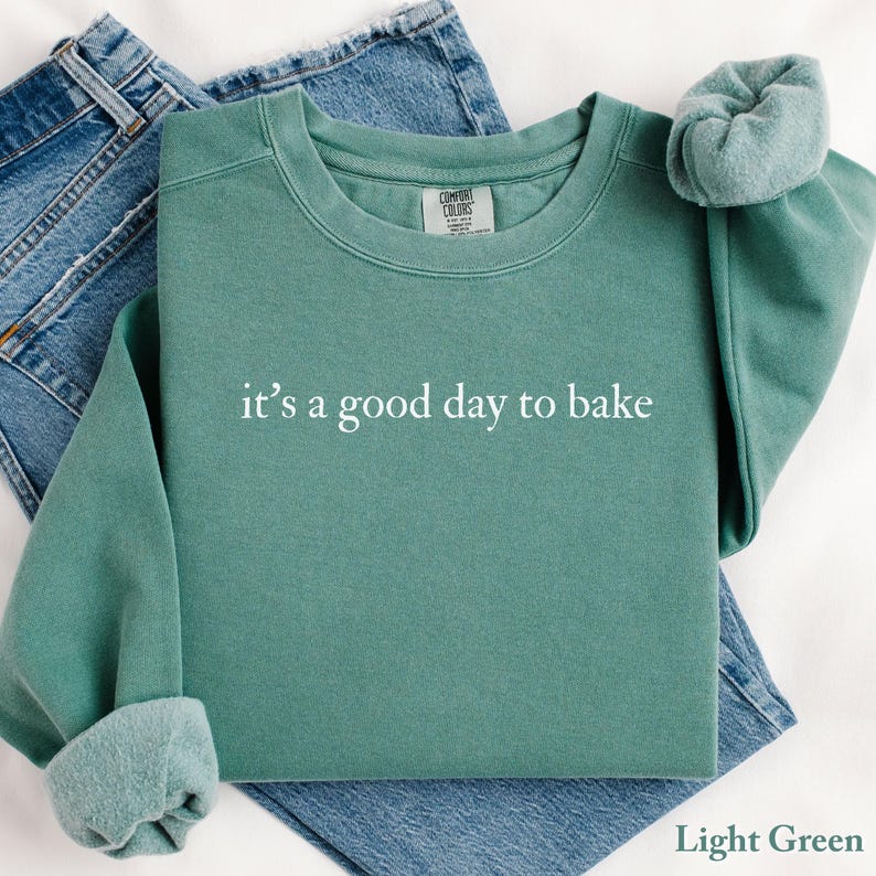 Puede incluir: Sudadera verde claro con el texto blanco "it's a good day to bake". La sudadera est&aacute; doblada sobre unos vaqueros azules. Las mangas est&aacute;n dobladas. Las palabras "Light Green" est&aacute;n en la parte inferior derecha.