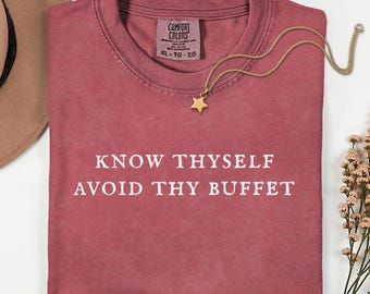 May include: A crimson crew neck t-shirt featuring the white text "KNOW THYSELF AVOID THY BUFFET" printed on the front. The shirt has a Comfort Colors label at the inner neckline and is displayed with a gold star pendant necklace.