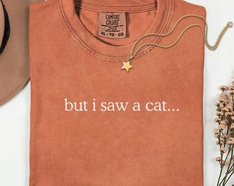 May include: A rust-colored short-sleeve t-shirt with a crew neckline and the white text "but i saw a cat..." printed on the front. The shirt features a Comfort Colors brand label at the inner neck and is paired with a gold-toned star pendant necklace.