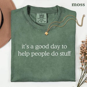 May include: A moss green crew neck t-shirt featuring the white text 'it's a good day to help people do stuff' printed on the front. The shirt has a Comfort Colors brand label at the inner neckline and is displayed with a gold star pendant necklace.