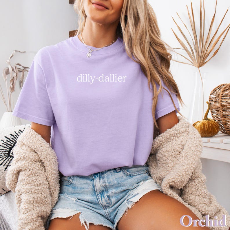 May include: A light purple t-shirt with the word "dilly-dallier" printed in white. The shirt is worn with denim shorts and a cream-colored cardigan. A necklace is visible. The word "Orchid" is at the bottom right.