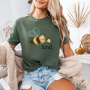 May include: Moss green t-shirt featuring a cartoon bee holding a flower, with the word "kind" printed below. The bee has yellow and black stripes and transparent wings. The shirt is a short-sleeved, casual style.