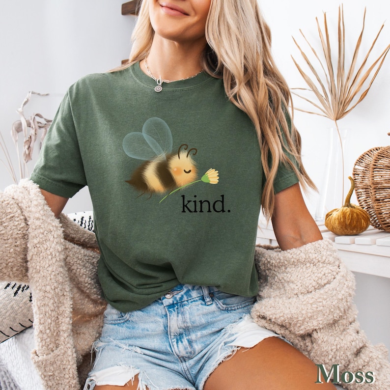 May include: Moss green t-shirt featuring a cartoon bee holding a flower, with the word "kind" printed below. The bee has yellow and black stripes and transparent wings. The shirt is a short-sleeved, casual style.