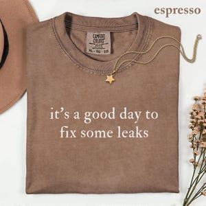 May include: An espresso-colored t-shirt with the white text 'it's a good day to fix some leaks' printed on the front. The shirt features a Comfort Colors label at the inner neckline, a crew neck, and short sleeves, presented in a flat lay style.