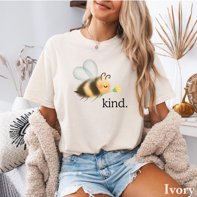 May include: Ivory-colored t-shirt featuring a cartoon bee holding a flower, with the word "kind" printed below. The bee has yellow and black stripes, with blue wings. The shirt is a relaxed fit.