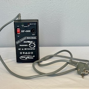 Puede incluir: Convertidor AC a AC negro, modelo SF-500, con cable de alimentación gris. El dispositivo tiene un interruptor rojo y muestra "500Watt" y "Automatic". El cable tiene un enchufe europeo.