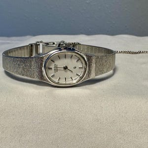 May include: A silver-tone Citizen Quartz wristwatch featuring an oval-shaped white dial with thin black hour markers and hands. The watch has a textured, brushed metal link bracelet and a matching silver-tone metal safety chain attached to the clasp.